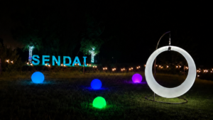 Night outdoor art installation in Sendai with glowing blue, green, and purple spheres on the lawn and a large illuminated circular sculpture on the right.